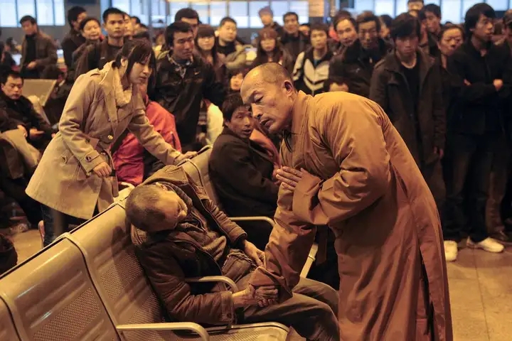 图片中,一位身着棕色僧袍的僧人正俯身握住坐在椅子上男子的手,神情专注。一旁还有一位穿着浅色风衣的女子似乎在关注着男子。背景中是一群聚集的人,场面显得较为拥挤。图片与上下文“佛家安宁”相关,可能意在体现佛教相关人士在安宁疗护等场景中给予关怀与慰藉的情景,展现了在生死教育与安宁疗护中的一种人文关怀画面。