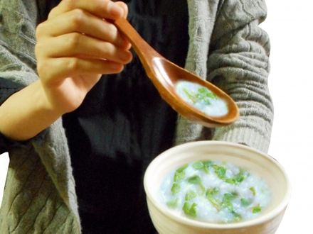 图片展示了一位穿着灰色毛衣的人端着一碗粥,用木勺舀起粥,勺中盛有粥和青菜。图片对应文档中“检查工作人员护理和护理的推荐时间是用餐时间”这一内容,特别是强调膳食援助是了解设施护理水平和工作人员对待居民情况的好机会,通过观察餐具材质、从轮椅转移到吃饭用椅子的行为等,可判断设施是否考虑安全、重视维护尊严生活。
